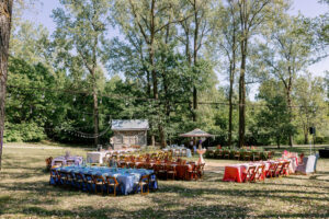 A colorful outdoor wedding reception in mid-Missouri with color-blocked table linens, wooden chairs, string lights, and vintage bud vases and candlesticks beneath tall trees.”